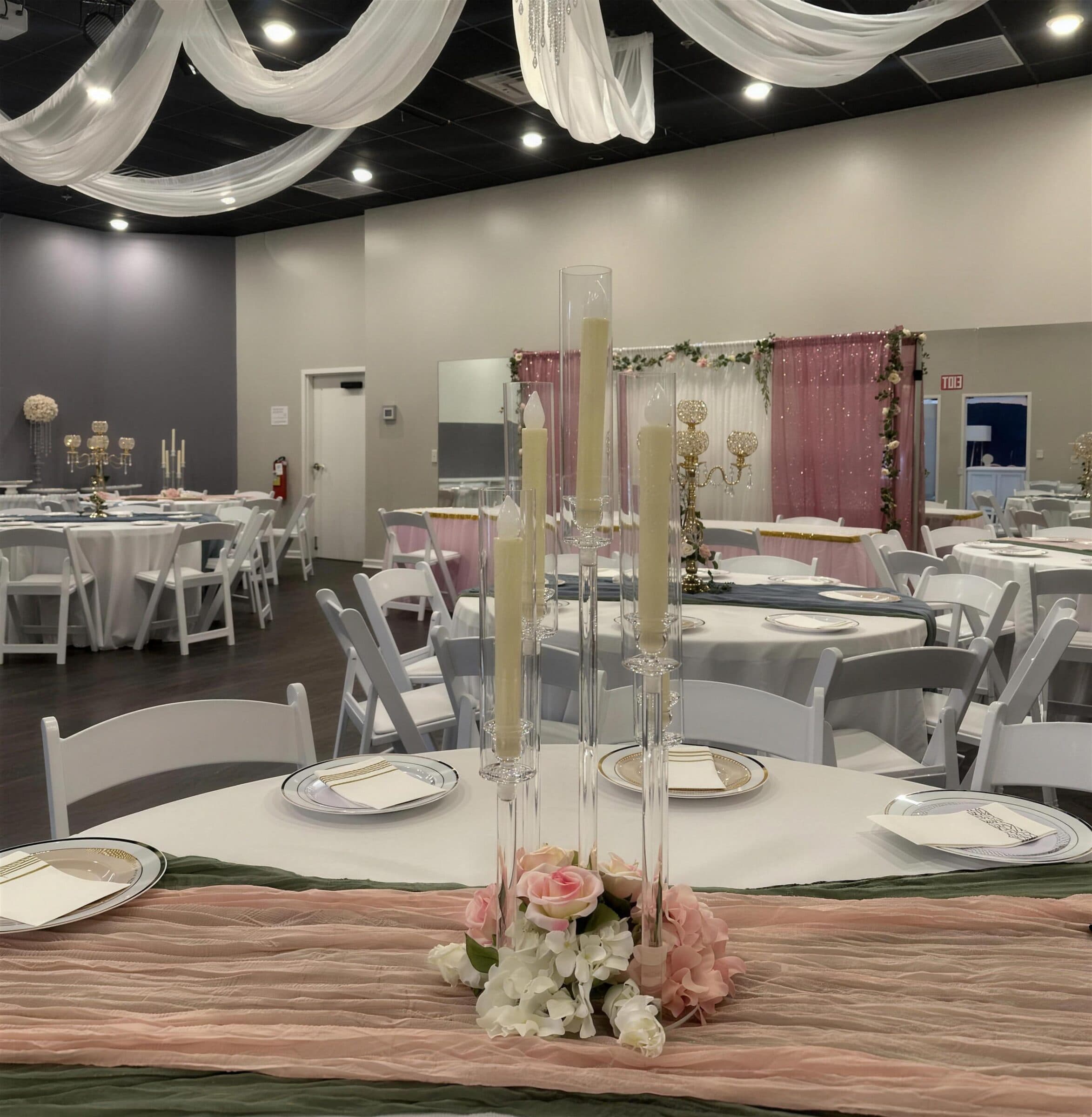 Round table set with silver-rim chargers, blush and sage runners and tall glass candlesticks rising from pink roses, blush sequin backdrop beyond, at The Cove at St. Johns, Fruit Cove, St. Johns County FL