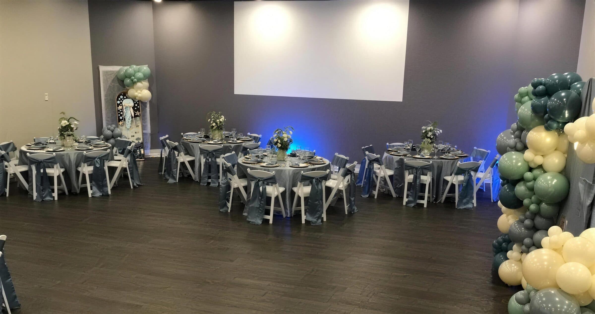 Dusty blue satin rounds and white folding chairs with a sage-and-cream balloon column beside a jellyfish art panel at a birthday celebration at The Cove at St. Johns, Fruit Cove FL