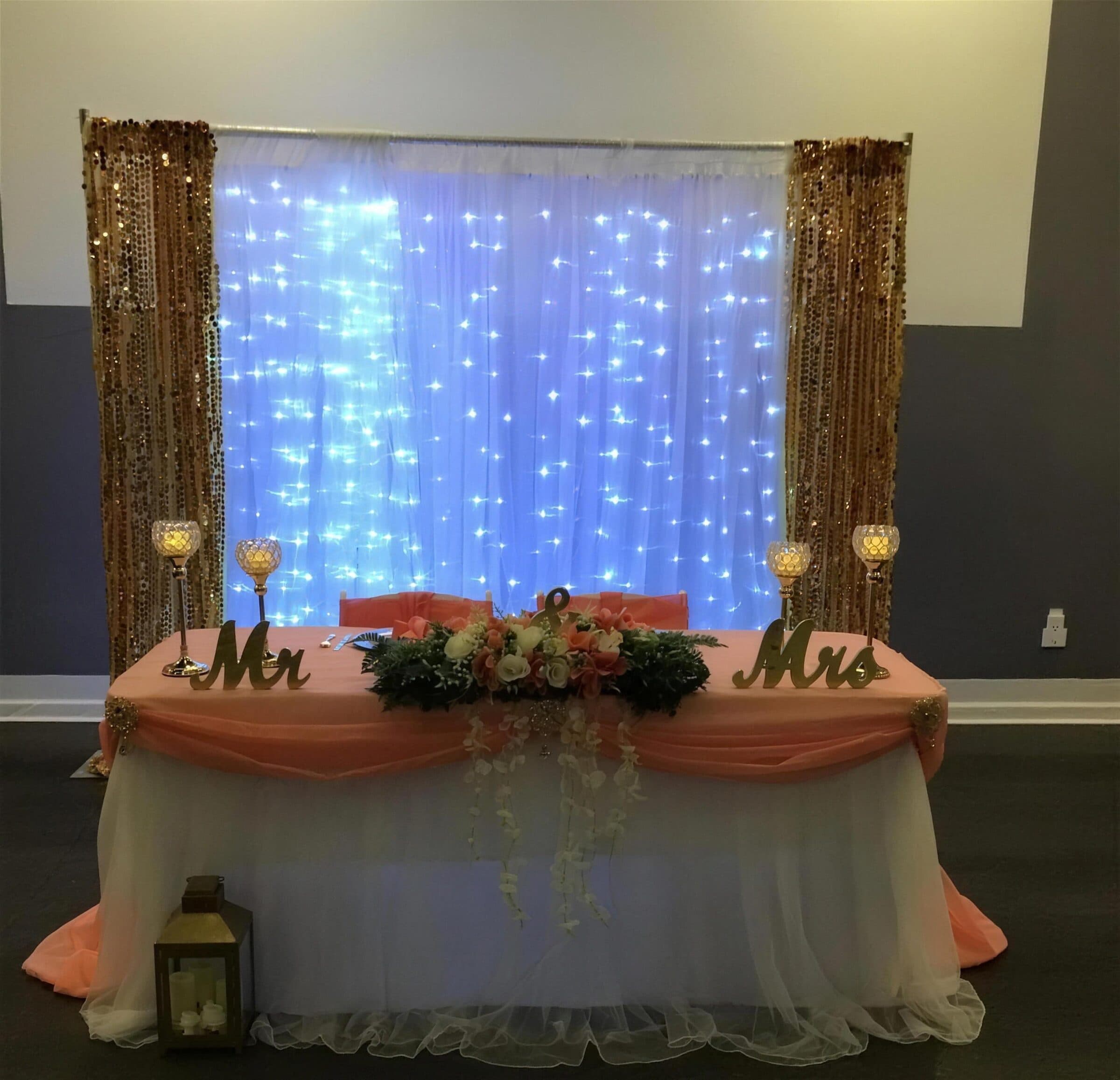 Peach organza and white tulle sweetheart table with gold Mr and Mrs letters in front of a fairy-light curtain framed by gold sequin panels at The Cove at St. Johns, Fruit Cove FL