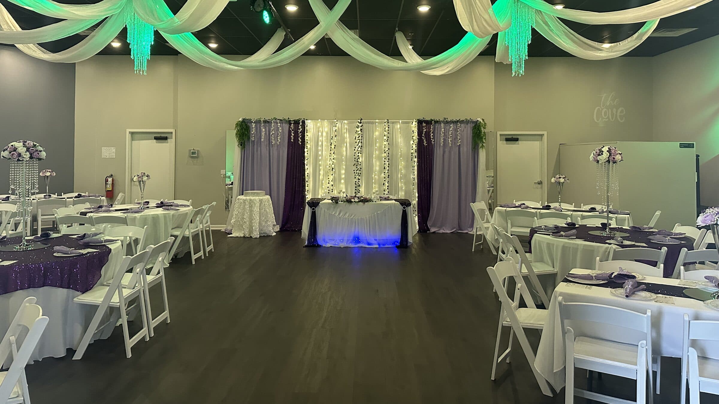 The same plum and lavender room from farther back with the ceiling drapes washed in teal light and the sweetheart table glowing blue at The Cove at St. Johns, Fruit Cove, St. Johns County FL