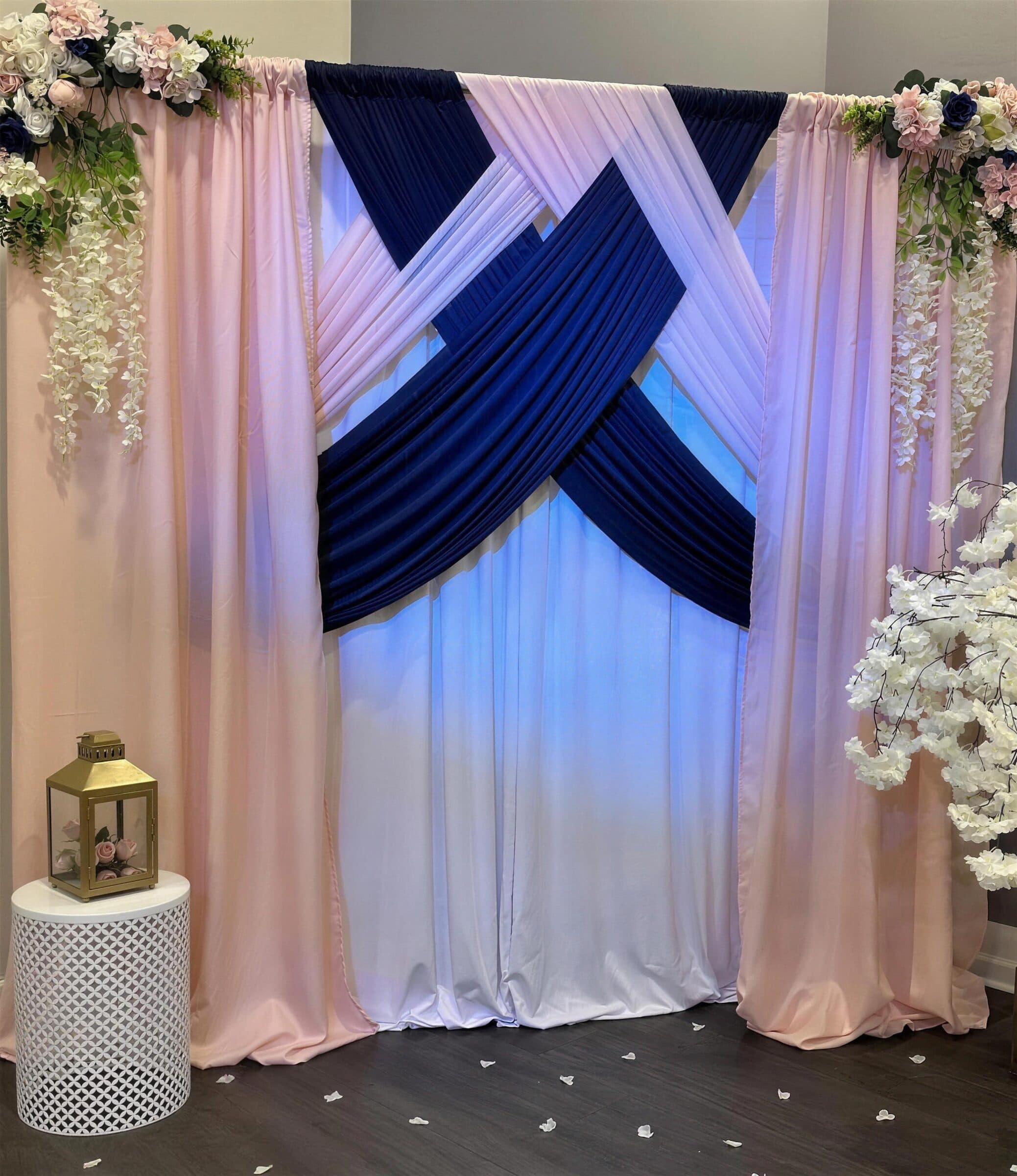 Blush and navy criss-cross drape backdrop trimmed with rose and wisteria swags, a gold lantern on a white side table and petals scattered on the floor at The Cove at St. Johns, St. Johns County FL